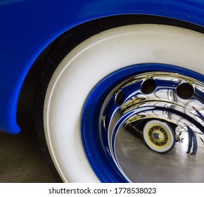 Wheel of blue vintage car with white tyre and shiny hubcap. Reflection in hubcap showing second car's wheel with white tyre and yellow spokes.