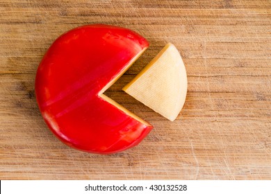 Wheel Of Fresh Gouda Cheese With A Red Rind And A Single Wedge Portion Cut Out, Peeled And Separated To The Side On A Wooden Cutting Board