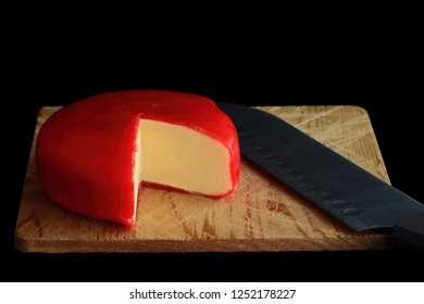 Wheel of Gouda Cheese covered with red wax protective layer over wooden cutting board, knife with which is cut a segment (wedge) of the cheese in order cheese to be seen over black background 