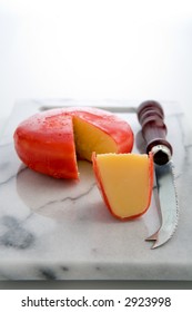 A Wheel Of Gouda Cheese Covered With Red Wax, With A Wedge Cut Out Form The Wheel