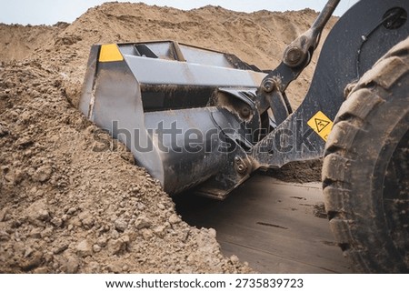 Wheel loader bucket scooping sandy soil while moving a large mound during groundwork, close side view of heavy earthmoving equipment at a dusty construction site, daytime progress