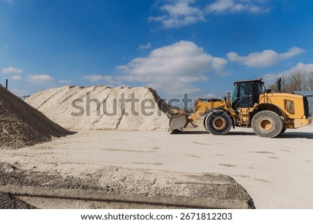 Wheel loader transporting sand within a concrete production plant, set against a backdrop of a bright blue sky dotted with clouds