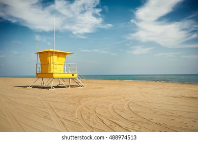 Wheel tracks curving on the sand next to an empty lifeguard post on an empty beach under dramatic blue sky with white clouds