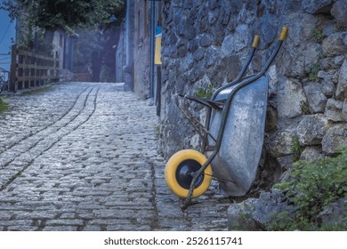 A wheelbarrow is sitting on a cobblestone path next to a stone wall. The wheelbarrow is old and rusted, and it is abandoned. - Powered by Shutterstock