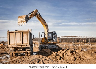 A wheeled excavator loads a dump truck with soil and sand. The excavator digs the ground at the construction site. Removal of soil from a construction site or quarry - Shutterstock ID 2271913931