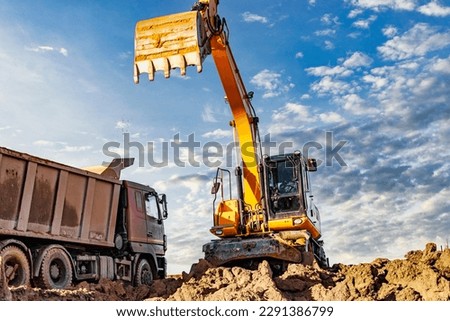 A wheeled excavator loads a dump truck with soil and sand. An excavator with a high-raised bucket against a cloudy sky View from the trench. Removal of soil from a construction site or quarry