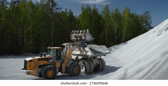 Wheeled Front-end Loader Loading Crushed Stone In Mining Dump Truck Against A Hill Of Crushed Stone And A Green Forest In Sunny Weather, Panorama. Mining Industry.