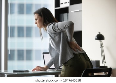 When Office Chair Is Uncomfortable. Sad Suffering Millennial Woman Employee Leaning Forward Above Work Desk Holding Hand On Lower Back Feeling Bad, Sudden Pain Attack, Trying To Massage Tensed Muscles