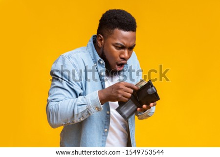 Where's my money. Shocked african american guy holding wallet with dollar cash that will be not enough to cover spendings, yellow studio background.