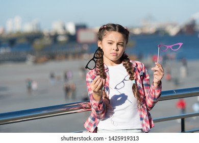 Which Props To Choose. Doubting Small Girl Choosing Glasses Props. Adorable Little Child Holding Photobooth Props On Sticks. Cute Kid With Fancy Party Props On Urban Outdoor.