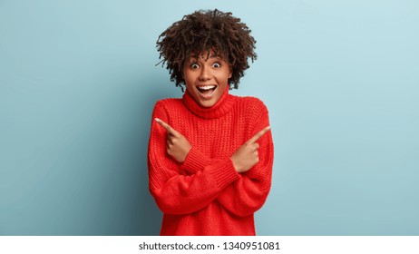 Which way way to choose? Cheerful lovely dark skinned woman points at different corners, has satisfied expression, wears oversized jumper, stands against blue studio wall. Body language concept - Shutterstock ID 1340951081