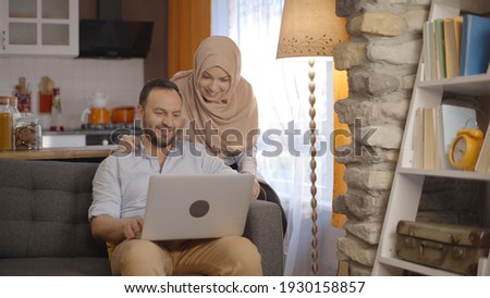 While the bearded man orders something from the computer, the young woman in the hijab tells his opinion to his wife. On line shopping concept.
