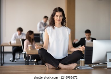 While diverse colleagues working, attractive young employee in coworking area sitting on office table in lotus pose and practising yoga meditating folding fingers in Jnana Mudra. Stress relief concept - Powered by Shutterstock