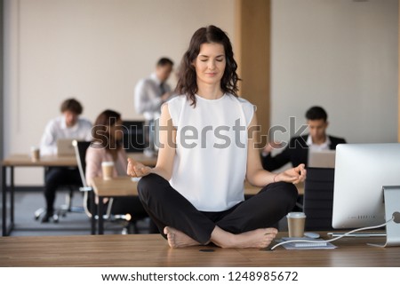 While diverse colleagues working, attractive young employee in coworking area sitting on office table in lotus pose and practising yoga meditating folding fingers in Jnana Mudra. Stress relief concept