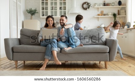 While funny active daughter and son running parents sit on sofa resting using pc online services plan family holiday choose tour booking online apartments, e-commerce usage on weekend at home concept