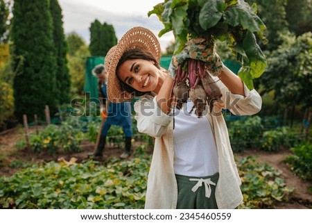 While her father works in the background, a woman holding up her beet and displaying them to the camera while smiling and wearing colorful gardening gloves