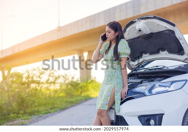 While looking at a broken down car on the street,\
a young woman is on her phone.\
Call an auto repair using a mobile\
phone when the car is\
broken.