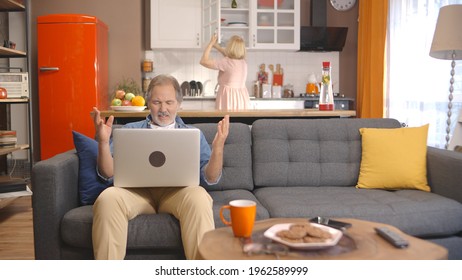 While The Old Man Working At Home From The Computer Gets Bored And Angry When He Makes A Mistake, His Wife Makes Something In The Kitchen. False Mailing Concept.