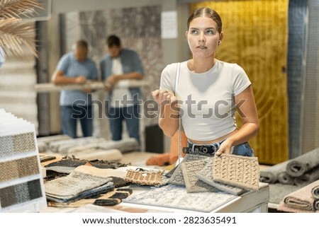 While visiting store with household and interior goods, girl examines examples of carpet products, chooses carpet material. She inspect small pieces of carpet, make decision about purchase.