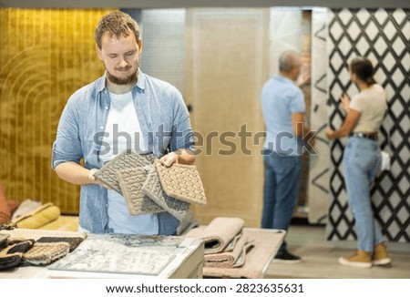 While visiting store with household and interior goods, guy examines examples of carpet products, chooses carpet material. He inspect small pieces of carpet, make decision about purchase.