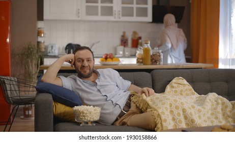 While watching something funny on TV with a blanket on the sofa, the bearded man eating popcorn laughs a lot. 