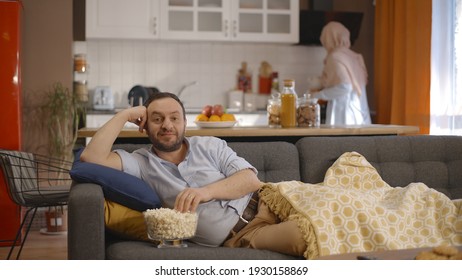 While watching something funny on TV with a blanket on the sofa, the bearded man eating popcorn laughs a lot. 