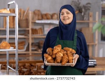 While working in bakery store, Asian Muslim woman in hijab and apron sells croissants, replenishes assortment in store window, shows goods
