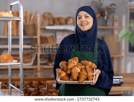 While working in bakery store, Asian Muslim woman in hijab and apron sells croissants, replenishes assortment in store window, shows goods