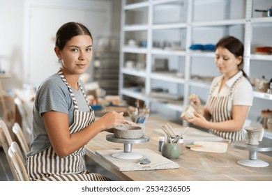 While working in pottery workshop, girls colleagues produce ceramic decorative products. Young worker makes blank from raw clay, apprentice paints product with paints - Powered by Shutterstock