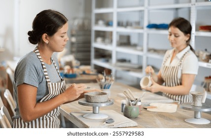 While working in pottery workshop, girls colleagues produce ceramic decorative products. Young worker makes blank from raw clay, apprentice paints product with paints - Powered by Shutterstock