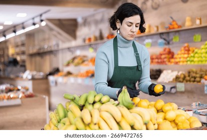 While working in shop, employee forms display case, puts ripe imported lemon in pile. Open display of goods in self-service shop. Female employee makes attractive display case with vegetables.