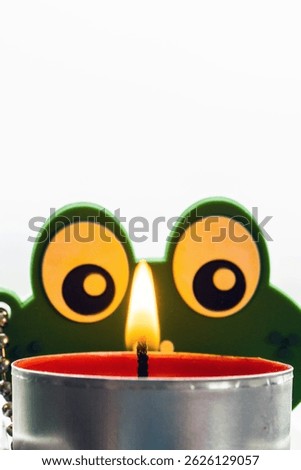 Whimsical Green Frog Eyes Decoration Peeking Over a Lit Tealight Candle Flame, Fun Still Life