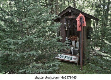 A whimsical outhouse set in among a thick forest of trees. A humorous office sign, life jacket, and other fun items have been added for a good laugh. Horizontal format for outside the box thinking