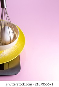 Whisk with an egg inside, placed in sieve with flour, in plate that stands on the scales. Pink background with space for text. Picture for websites about food, baking, confectionery. Selective focus.
