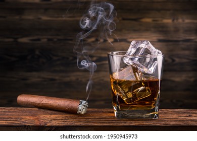 Whiskey and cigar. Glass of whiskey with smoke cigar. Bourbon or Brandy Glass with ice cube and cigars from Cuba Havana on natural wooden background. Alcohol drink on Bar counter in the restaurant. 