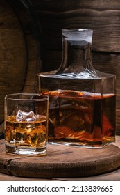 Whiskey With Ice, Vertical Format. Decanter Of Old Whiskey And Whiskey In Glass Of Ice Against Backdrop Of Old Barrel Of Full Alcoholic Beverage On Wooden Background In Old Basement.