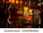 Whiskey is poured from a bottle into a glass with ice. Glass of whiskey on a black reflective background. In the background bottles with various alcoholic drinks.