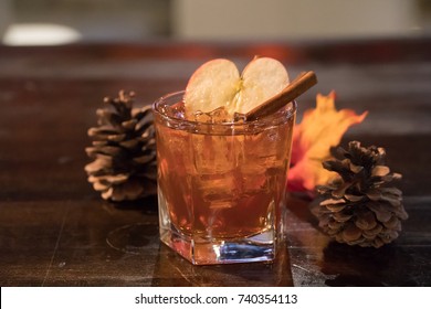 Whiskey Rum Cocktail Fall Beverage In Bar Lounge With Autumn Leaves, Pinecone, Apple, And Cinnamon Stick