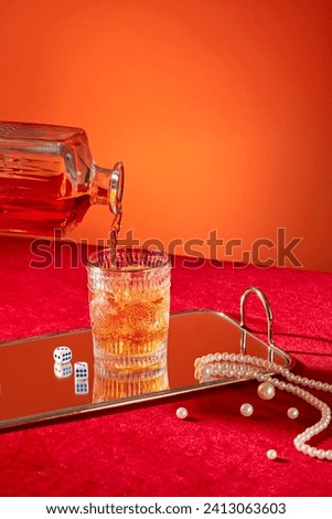 Whisky is poured into a glass from a decanter. Front view, whisky cup displayed on mirror sheet with dices and pearl necklace on orange and red background. Space for design