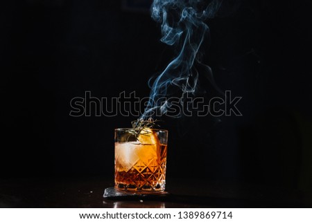 Whisky & Smoke. Smoking whiskey with ice and orange on a dark background. Cognac with smoking branch of lavender. Experimental alcoholic cocktail in a glass with smoking rosemary.