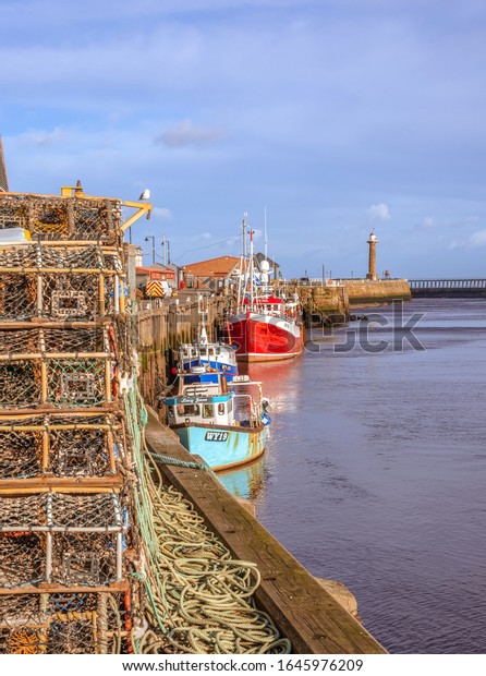 Whitby, UK.  February 11,\
2010.  Whitby harbour with three boats moored alongside a wharf. \
Lobster pots are the in the foreground  and a lighthouse is in the\
distance.