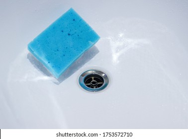 A White Acrylic Bathtub With A Metal Chrome Sewer Drain In Water Drops And A Blue Sponge For Cleaning The Tub. Selective Focus. Cleaning And Disinfection Of The Bath And Shower.