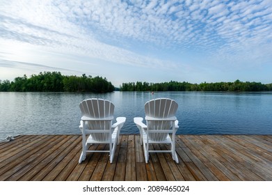 White Adirondack chair on a wooden dock facing the blue water of a lake in Ontario Canada. Cottages nestled among green trees are visible across the water. A single scull rower is passing.