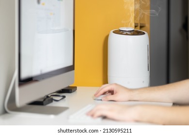 White Air Humidifier Spreading Steam Up. Humidification Of Dry Air While Man Working On Computer. Selective Focus On Vapor. Air Purity And Health Care Concept. Increasing The Humidity In The Room.