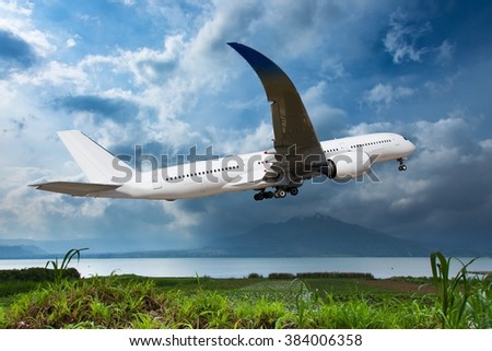 White aircraft is climbing in the blue sky. The plane flies over the lake, which shore overgrown with green grass. And the high mountain at the background.