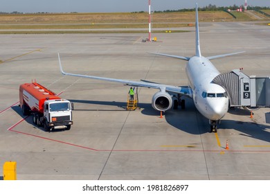 White Airplane Docked On Apron Outside Airport Terminal With Jet Bridge. Worker Refuels Plane. Fuel Hose From Red Truck Connected Under The Fuel Tank In The Wing. Ground Service Theme.