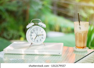 White Alarm Clock, Laptop And Iced Coffee Mug On Desk With Beautiful Bokeh Blurred Nature For Background. Time To Start With New Beginning.