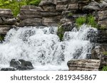 A white artificial waterfall flows down between cascading gray rocks, surrounded by lush garden plants.