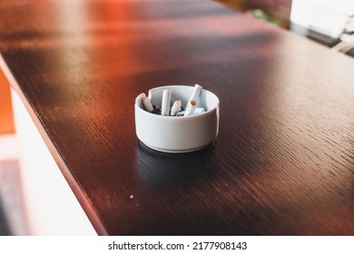 White Ashtray Full Of Cigarette Butts Or Stubs On A Wooden Bar Table