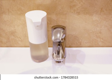 White Automatic Touchless Foam Foap Dispenser In Bathroom Standing On White Bathroom Sink Near To Chrome Faucet Against The Brown Tiled Wall Background.